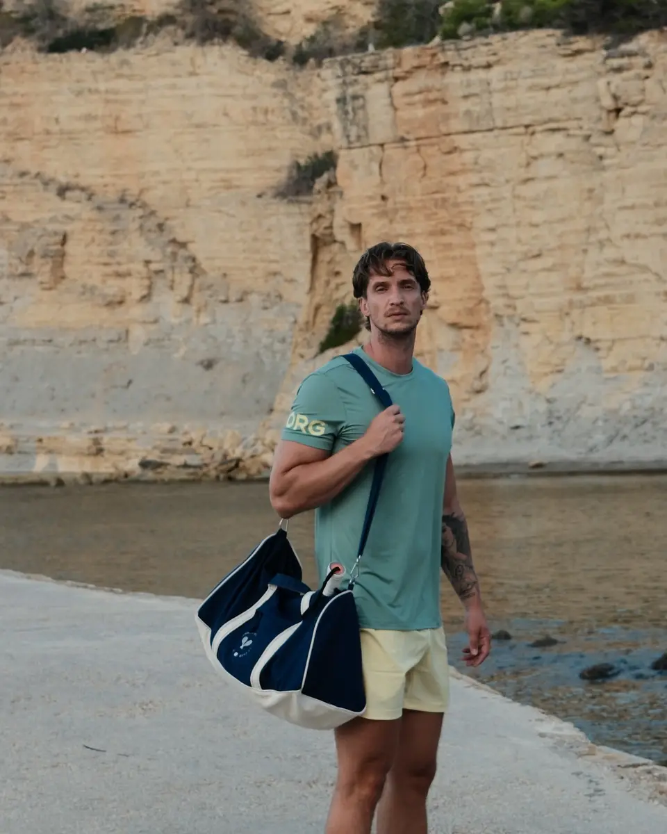 Arjan loopt over de kade met een marineblauwe sporttas over zijn schouder, in een groen Björn Borg-shirt en gele short