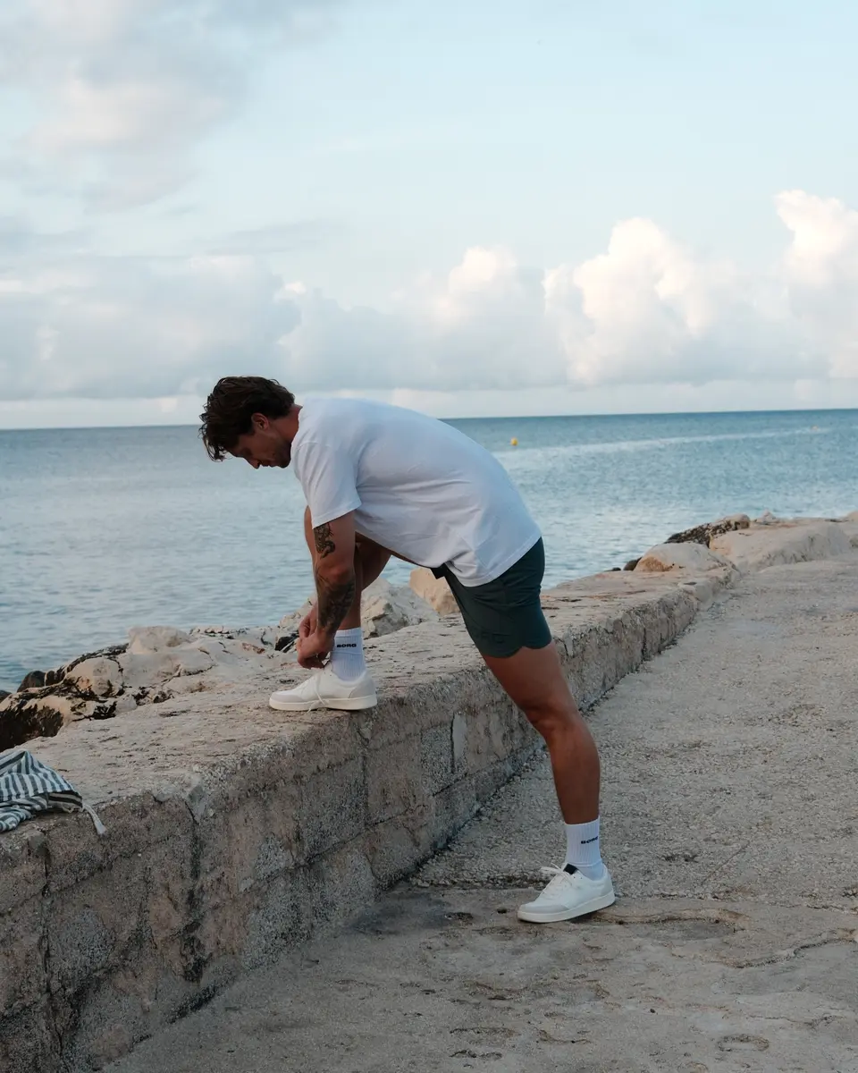 Arjan strikt zijn veter met zijn voet op een muurtje aan het water, in een wit oversized shirt