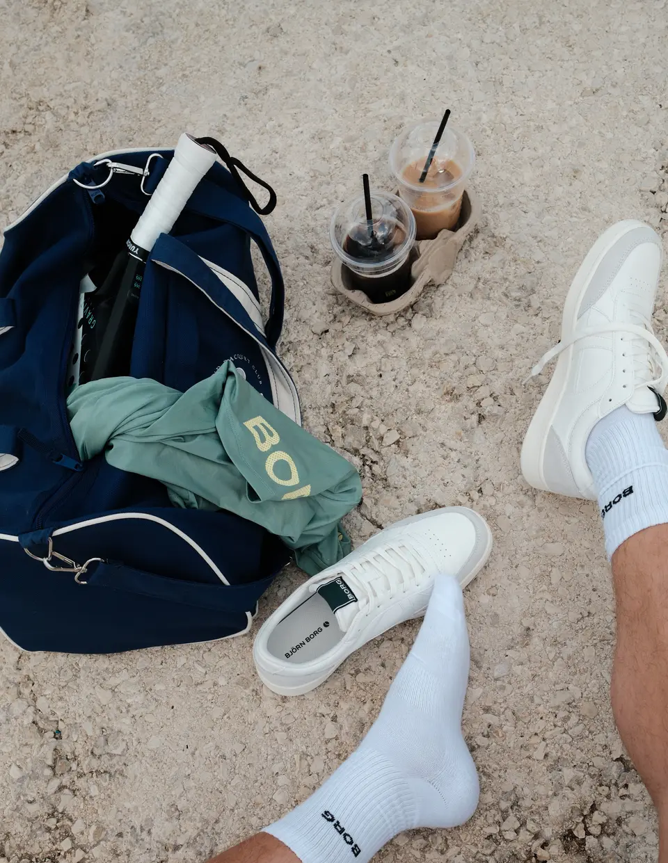 Van bovenaf: de open sporttas met een racket en een groen Björn Borg-shirt, twee ijskoffies op het grind, en beide paar witte sneakers naast elkaar