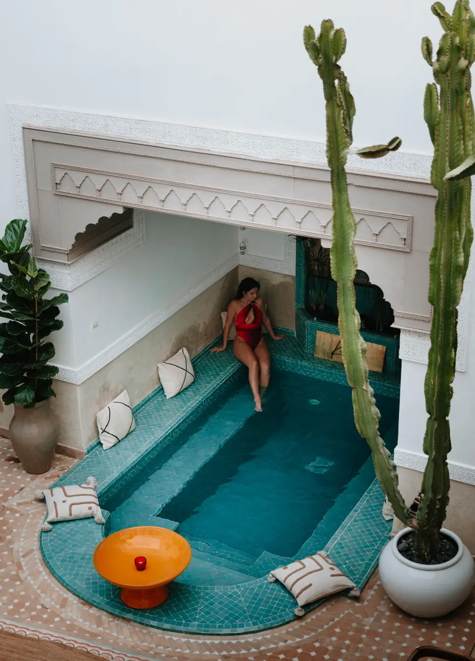 Nikki op de trap van een turquoise betegeld zwembad in een riad-binnenplaats, met een cactus en een oranje bijzettafel ernaast