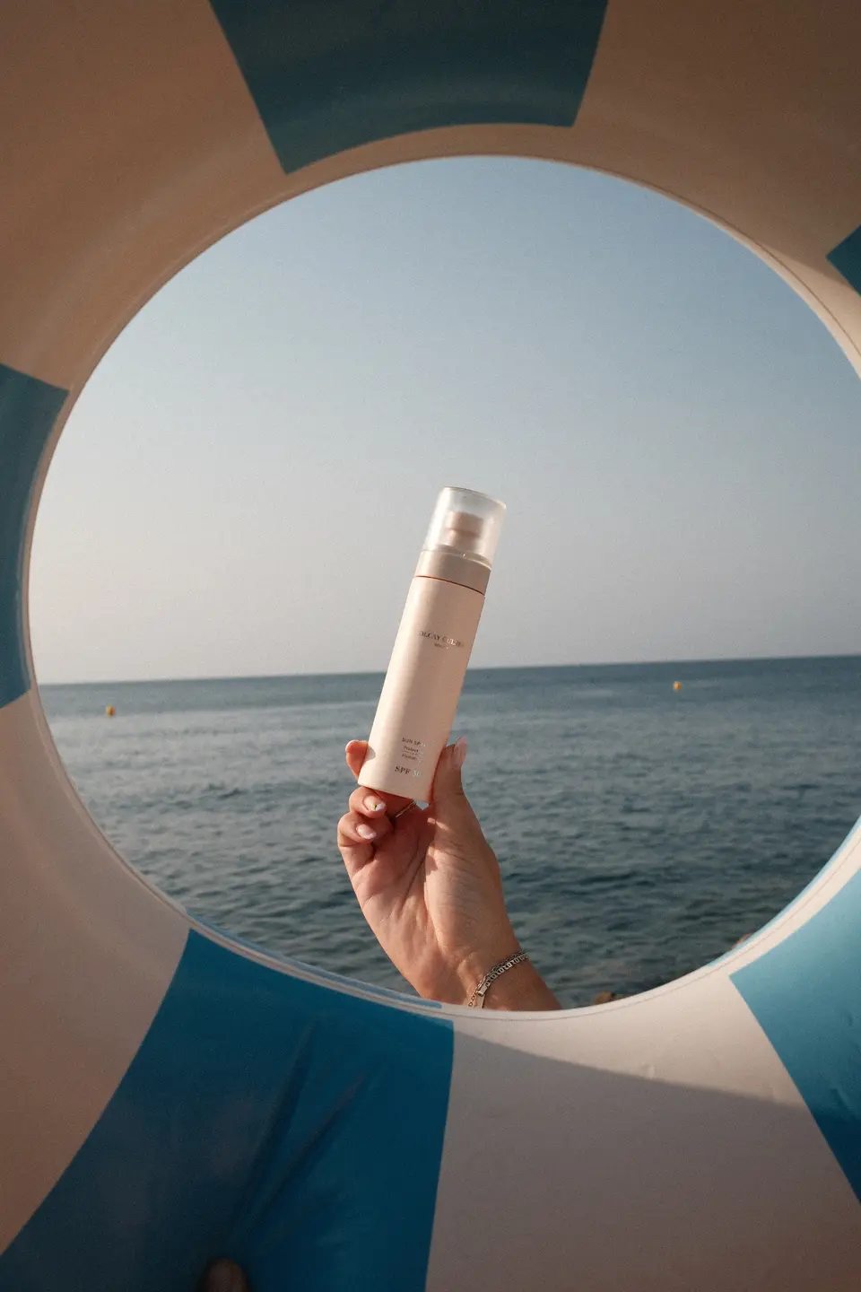 Door de opening van een blauw-wit gestreepte zwemband: een hand houdt een fles Olcay Gulsen Beauty Sun Spray SPF 50 omhoog tegen de zee en de horizon